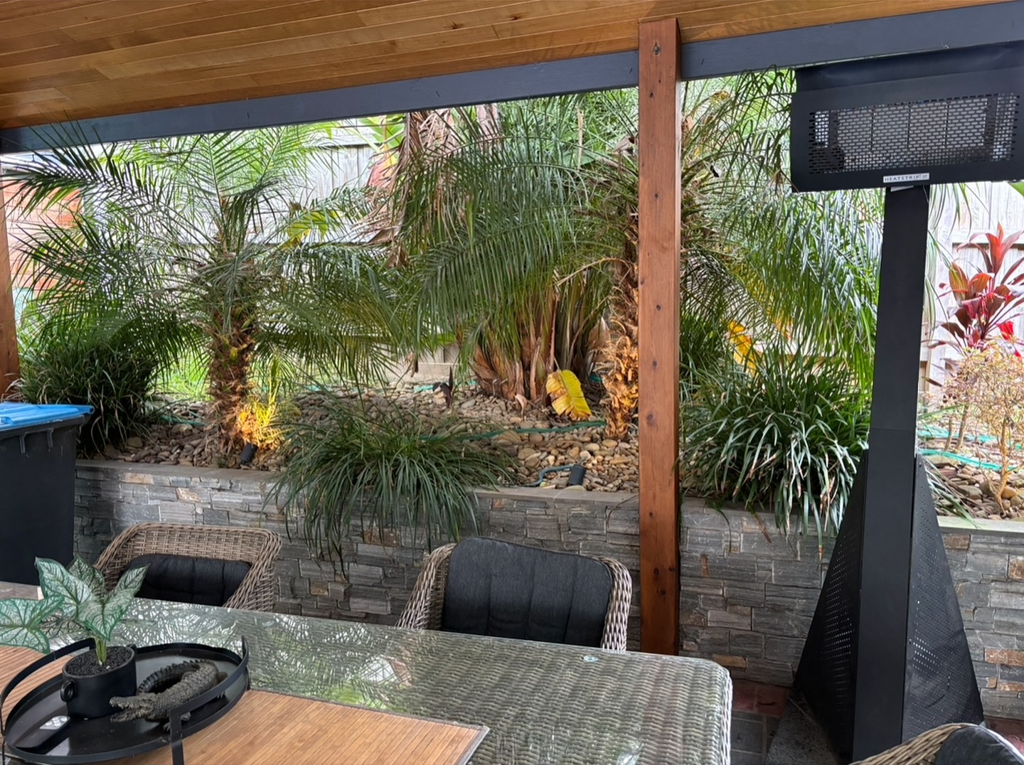 Covered alfresco dining area beside a stone-clad raised garden bed with palms