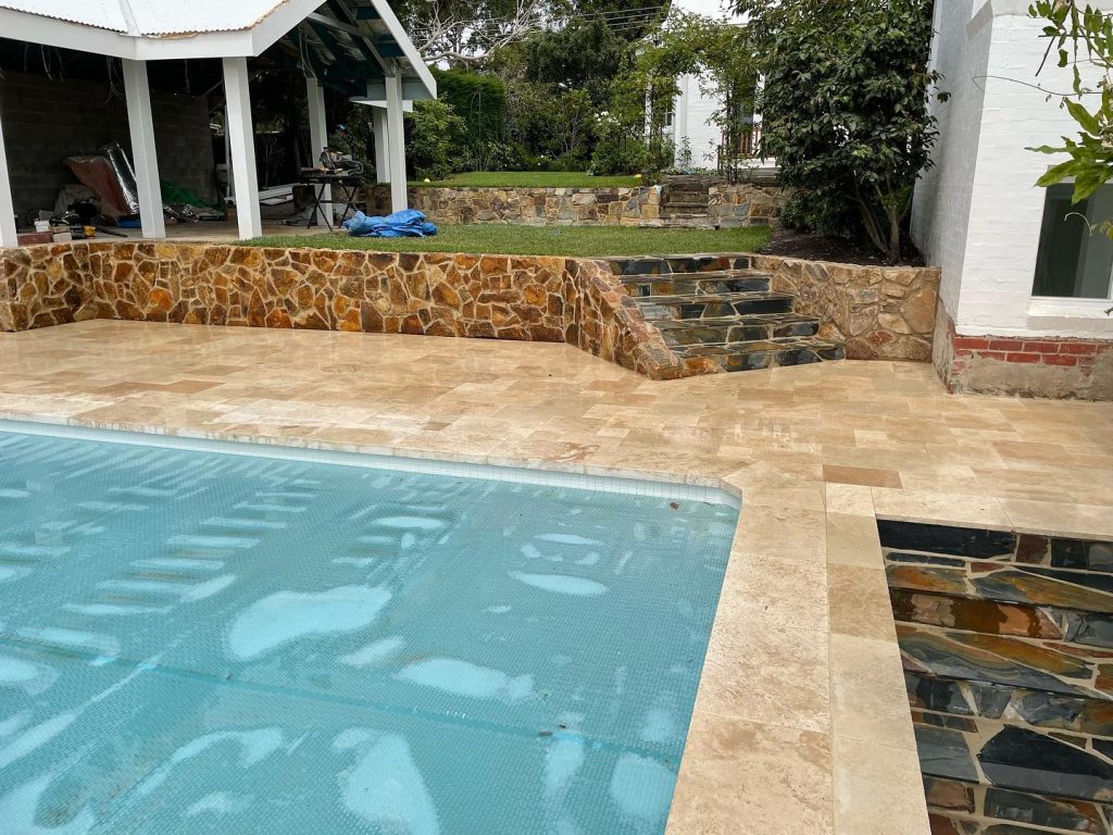 Pool surrounded by cream stone paving, with a stone retaining wall and steps behind